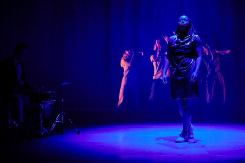 Ghaliah Conroy lit in blue centre stage with the rest of the ensemble in movement in the background