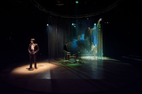 Alex O'Neill spotlit with drums and a figure dancing in darkness behind