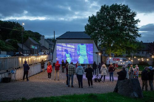Audience gathered around the opening of a handball alley with projections cast on its back wall