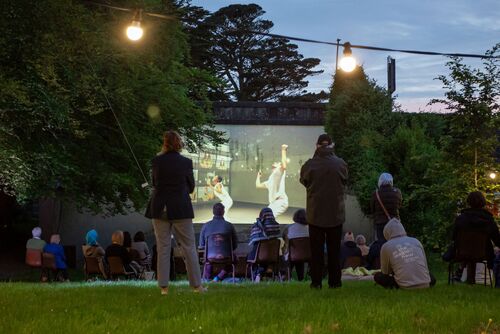 Audience gathered around the opening of a handball alley with projections cast on its back wall