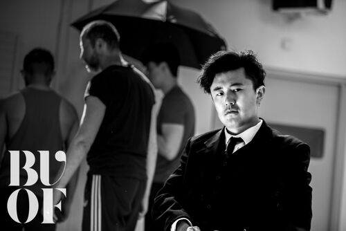 A seated figure with performers in movement in the background