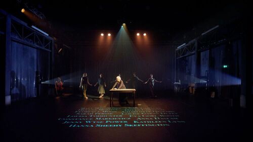 Máire Ní Ghráinne behind a table centre stage with dancers in movement behind