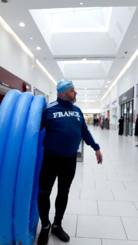 A man in a swim cap stands in a shopping centre with a blue inflatable pool