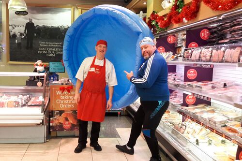 A man in a swim cap poses with a butcher in his shop holding an inflatable blue pool behind them