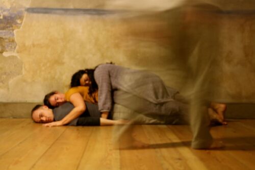 Three figures laid on top of each other on the floor with a pair of legs blurred in motion in front