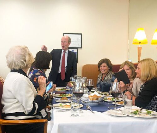 A figure in a suit addressing participants seated at a table set for a meal