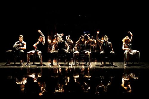 Six figures seated in a line on individual chairs in movement