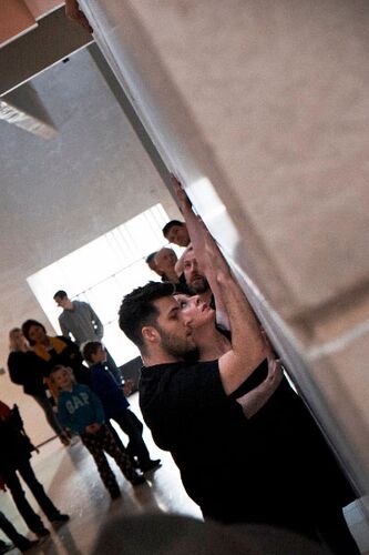 Two figures in movement against a wall seen in side profile with audience standing around them