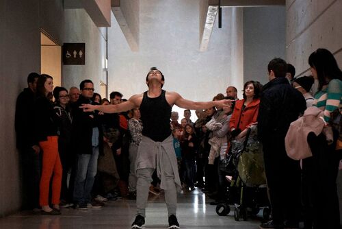 A figure in movement with arms outstretched surrounded by audience standing