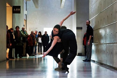 Two figures in movement together in a gallery space with audience watching in the background
