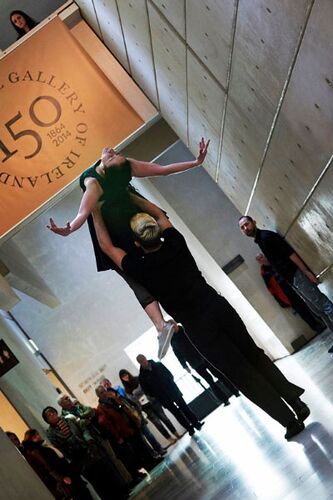 Two figures mid-lift in a gallery space with audience watching in the background