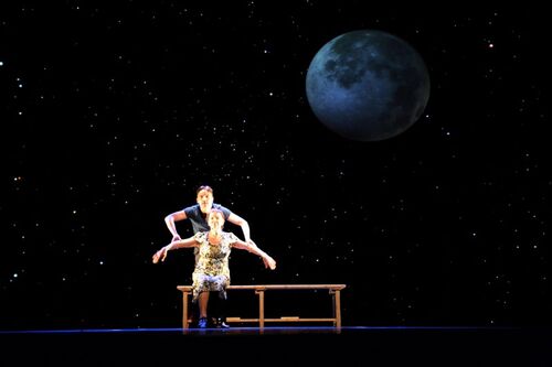 Two figures, one sat on a bench and the other bent over her from behind with a night sky backdrop