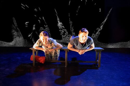 Two figures bent over a bench looking out to audience