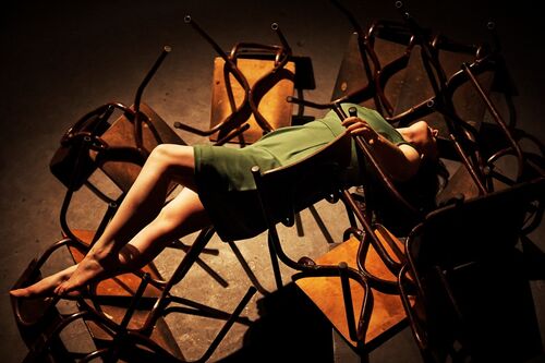 A figure draped across a chair surrounded by other upturned chairs