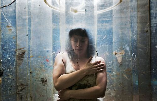 A figure with arms wrapped around her body behind a curtain of heavy plastic