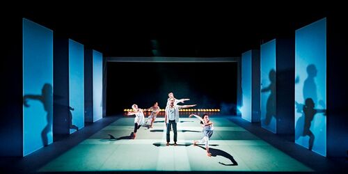 Figures in movement centre stage with shadows cast along large screens along the sides of the stage