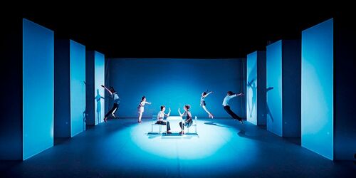 Two figures seated at a table centre stage with two figures in movement at either side