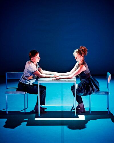 Two figures seated opposite each other at a table on a blue lit stage