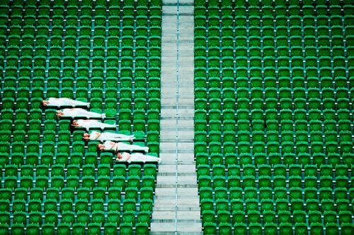 Six figures dressed in white lying staggered across rows of green stadium seats seen from afar