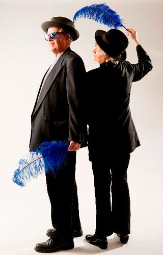 Two figures in black suits posing with bowler hats and blue feathers in hand