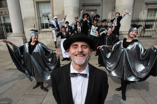 A participant smiling into the camera with an ensemble of others in full costume scattered behind