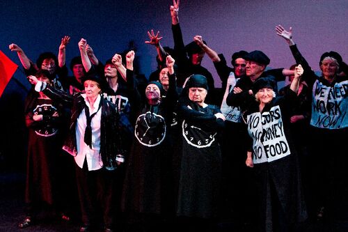 An ensemble of participants dressed in black with signs and clocks on their fronts on stage