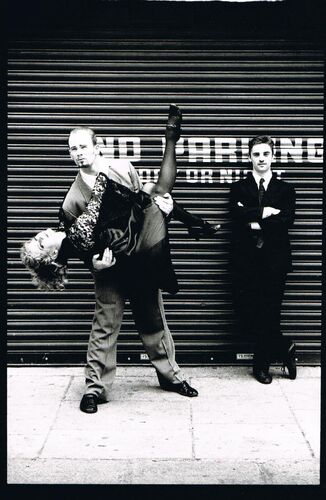 Three figures on the street in front of a shutter, two in movement mid-lift and the third standing