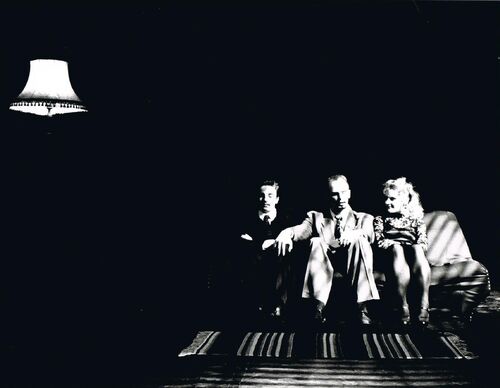 Three figures seated on a sofa with a lamp at the opposite side of the frame