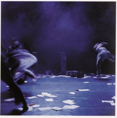 Three figures blurred in motion on a stage littered with sheets of paper