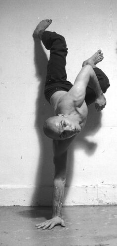 A figure in a one-handed handstand against a wall looking to camera