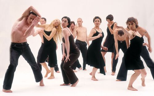 An ensemble of dancers dressed in black in movement on a white backdrop