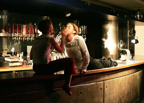 Two figures on top of a bar counter - one seated feeding the other lying on their stomach