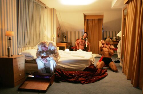 Two pairs in movement in a hotel room with an older figure on the bed looking into a suitcase