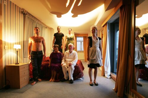 Two older figures seated on a bed and four others standing around the bed in a hotel room