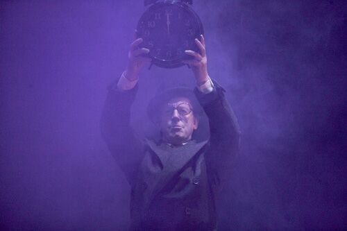 A figure holding a large clock aloft on a hazel filled stage