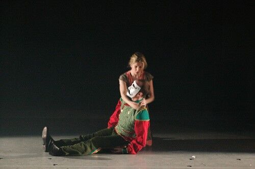 A figure in a mask being held in the arms of another figure on the stage floor