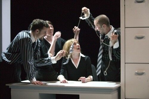 An ensemble dressed in office attire gathered around a desk and filing cabinet