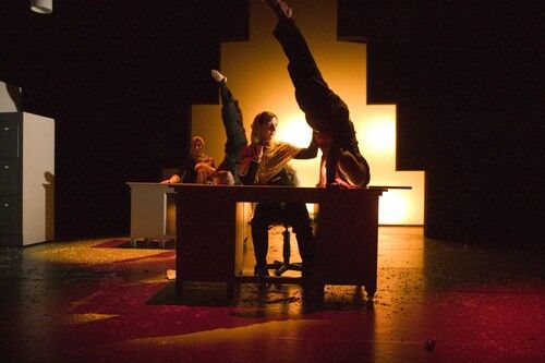 A figure seated at a desk with another in a headstand, their head disappearing into the desktop