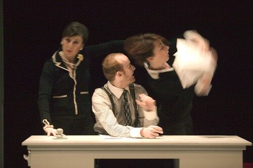 Three figures in movement with one seated at a desk and the other two standing at either side