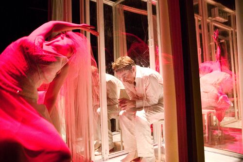A figure seated hunched in a perspex cubicle with another figure cast in pink in movement outside