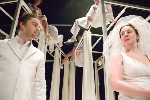 Two figures in all white formal attire with an ensemble sitting atop large frames behind them