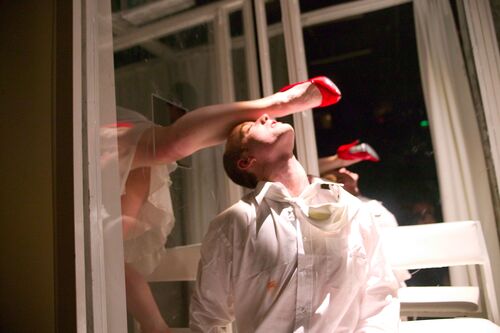 A red stiletto clad leg coming through a perspex wall and resting on a dancer's upturned face