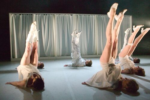 An ensemble of dancers in movement on the floor all with legs raised straight in the air