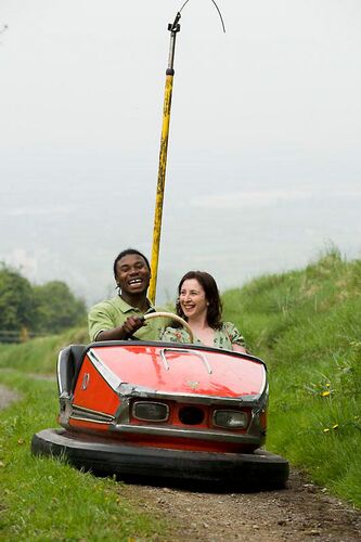 Two figures seated in a dodgem car on an unpaved path through grass