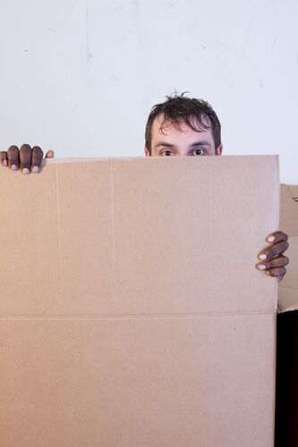 A pair of eyes peeking over a large square of cardboard with another's hands gripping either side