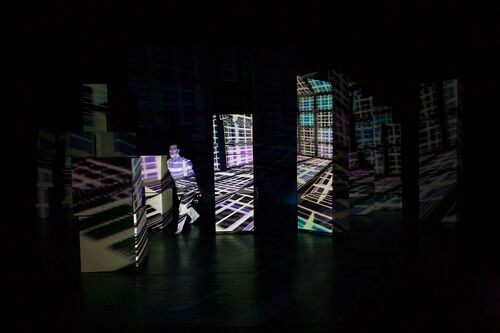 A figure among tall stacks of cardboard boxes with projections cast on them