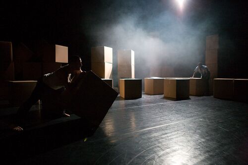A stage scattered with cardboard boxes and two figures moving among them