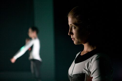 A close up of a figure in side profile on stage with another figure in movement in the background