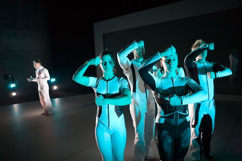 Four figures lit in blue in movement near the camera with another figure standing in the background