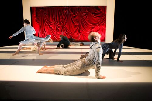 An ensemble staggered upstage in movement on the floor with one figure standing in movement